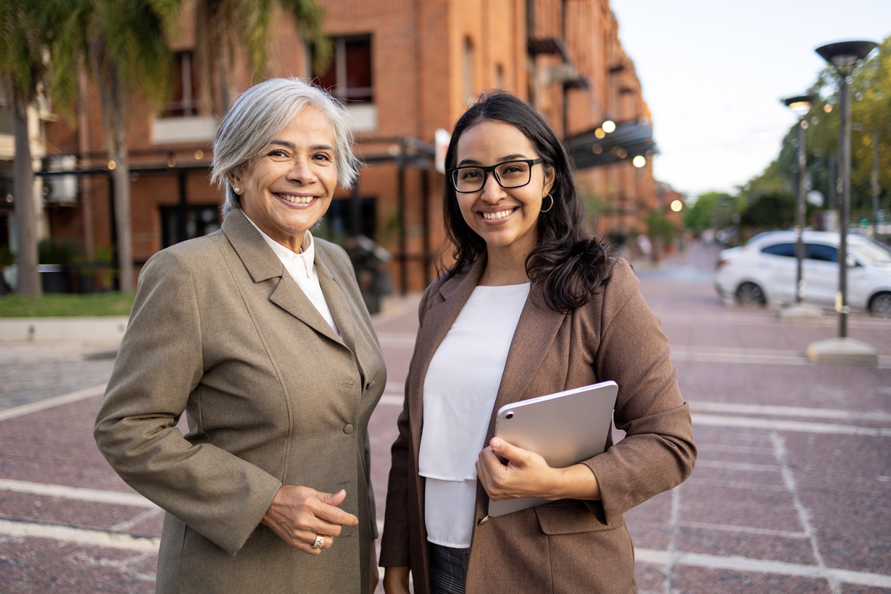 Two professional women standing outdoors in a business district downtown area. They are looking at the camera Business Succession Plan